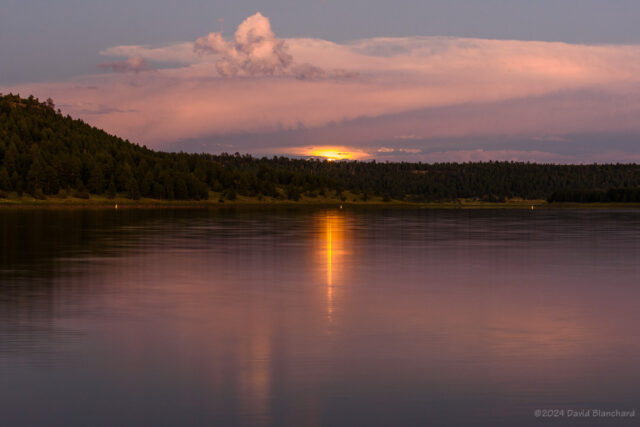 A small sliver of the rising Moon is visible in the gap in the clouds and casts a long reflection on the lake.