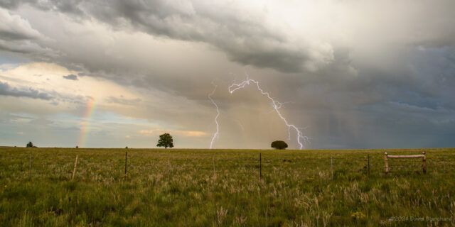 A lightning bolt with multiple landing points and a partial rainbow over the grasslands of Anderson Mesa.