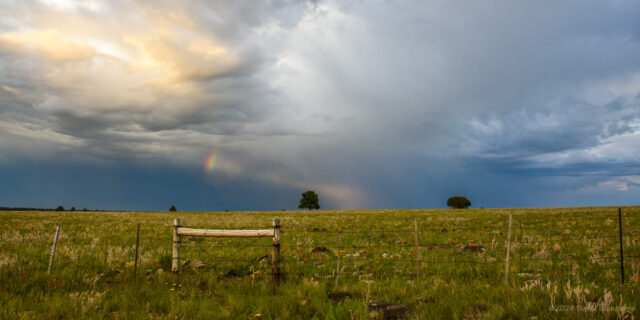 A beam of light pierces beneath a dissipating thunderstorm and creates a small segment of rainbow.